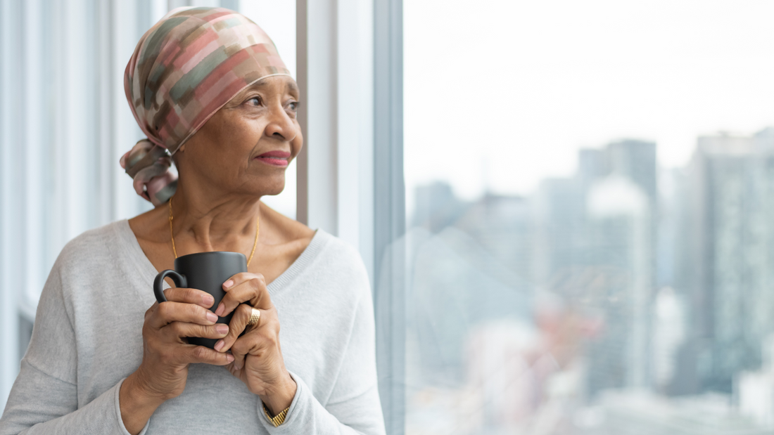 Older woman wearing headscarf holding a coffee mug and looking out a window, symbolizing cancer treatment and recovery support.