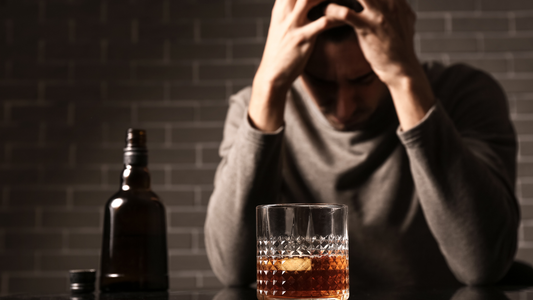 Man stressed with head in hands sitting near glass of whiskey and bottle symbolizing alcoholism disease crisis